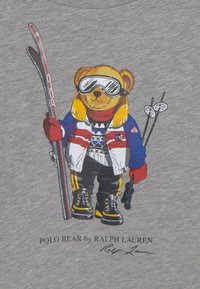 Miś pluszowy ubrany w strój narciarski, trzymający narty i kijki, noszący gogle, kurtkę, rękawiczki i buty na szarym tle.