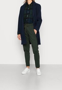 Marineblauer Mantel über einem dunkelgrünen karrierten Hemd, kombiniert mit grünen Hosen und weißen Sneakers. Der Mantel hat einen Reverskragen und eine Knopfverschluss.