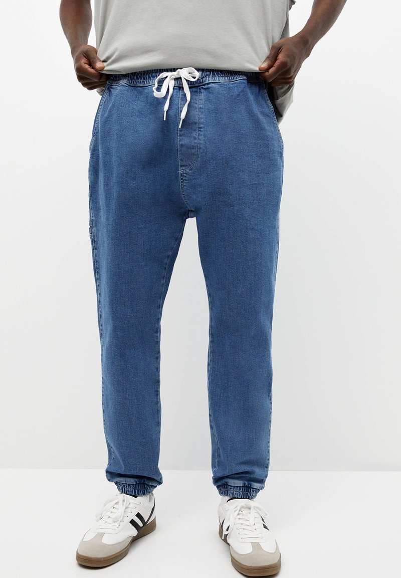 Man in blauwe denim joggers met elastische boorden en witte sneakers, die een lichtgrijs t-shirt aanpast.