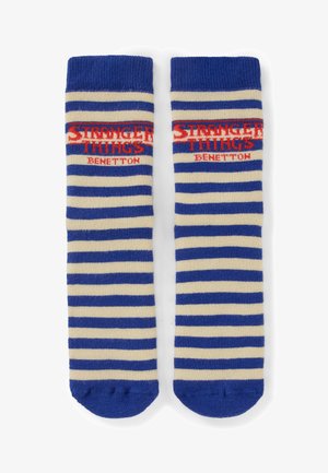 Blau-weiß gestreifte Socken mit rotem "Stranger Things Benetton"-Logo nahe dem oberen Rand, flach nebeneinander auf weißem Hintergrund gelegt.