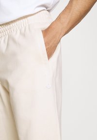 Handen i fickan på ljusbeige sweatpants med elastisk midja och en liten vit Adidas-logotyp.