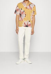 Korte mouwen, geel bloemenhemd met grote roze en zwarte bloemen, gecombineerd met witte broeken en witte sneakers. Casual ontwerp, comfortabele pasvorm.