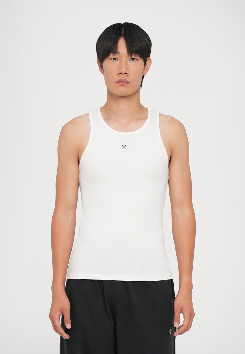 Hvid bomulds tanktop med brede stropper og en tætsiddende design, der har et lille logo på brystet. Båret med sorte, løstsiddende shorts.