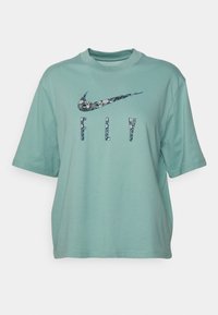 Mintvihreä ylimitoitettu T-paita, joka on valmistettu puuvillasta. Paita on varustettu sinisellä kuvioidulla Nike-logolla ja "FLY" -tekstillä, joka on myös kuvioitu. Lyhyet hihat.