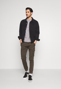Zwarte spijkerjas over een grijs T-shirt, in combinatie met olijfgroene cargobroeken met zijzakken, en zwarte sneakers met witte zolen.