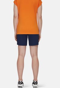 Orange kortärmad topp och marinblå shorts, båda tillverkade av lättviktsmaterial. Svarta och vita sneakers med ett mönstrat sula.