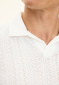 Biała dzianinowa koszulka polo z teksturowanym wzorem i geometrycznymi motywami. Kołnierzyk jest w jednolitym białym kolorze i kontrastuje z teksturą materiału.