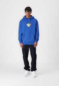 Blue hoodie with a front pocket and green graphic logo, worn with dark jeans and white sneakers; model is wearing sunglasses.