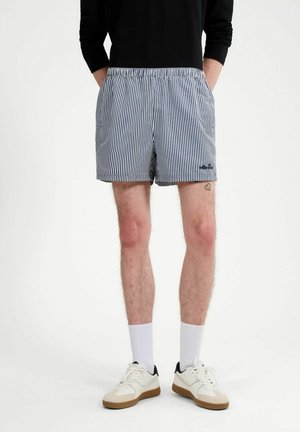 Hombre vestido con camiseta negra, pantalones cortos a rayas azules y blancas, calcetines blancos y zapatillas blancas con suelas de goma, de pie con las manos en los bolsillos.