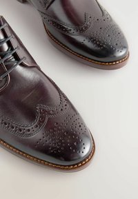 Tumed pruunid nahast brogues dekoratiivsete aukudega, terava ninaga ja seotud paeltega. Omab õmmeldud serva ja tekstuurset viimistlust.