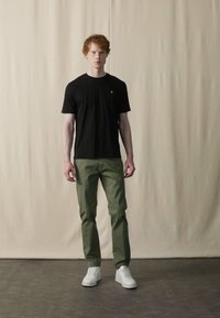Zwarte t-shirt met korte mouwen, groene broek en witte sneakers. Model staat tegen een neutrale achtergrond, met rechte benen en armen langs het lichaam.