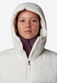 Vit pufferjacka med huva, med en lila fleecekrage och dragkedjedetalj. Vänster bröst har "The North Face"-logotyp.