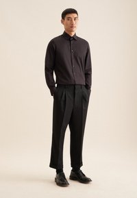 Zwarte lange mouwen shirt met een kraag en knopen, gecombineerd met brede zwarte broek en gepolijste zwarte schoenen, staande op een neutrale achtergrond.