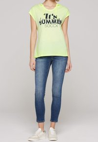 T-shirt à manches courtes à capuche de couleur jaune fluo avec l'impression "C'est L'ÉTÉ SOCCX", associée à un jean skinny bleu clair et des baskets blanches.