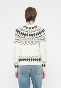 Oseba, oblečena v belo pulover s zelenimi in rožnatimi geometrijskimi vzorci, v kombinaciji z modrimi kavbojkami, prikazana s hrbtne strani na belem ozadju.