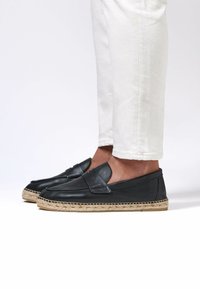 Zwarte leren espadrilles met een gladde textuur, een gestikte neus en een geweven jute zool, gedragen met witte omgeslagen broeken.