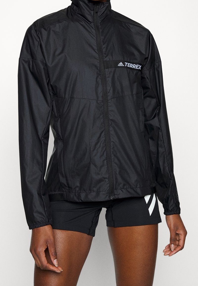 Chaqueta ligera con cremallera negra de Adidas Terrex, combinada con pantalones cortos deportivos negros que tienen franjas blancas, sobre un fondo liso.