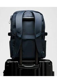 Jūrininkų kuprinė su paminkštintomis petnešomis ir reguliuojamais elementais, padėta ant juodo lagaminų rankenos, pasižyminti lygiu paviršiumi ir struktūrizuotu dizainu.