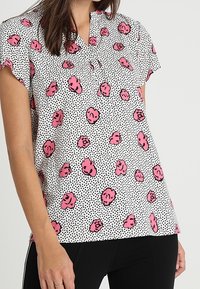 Blusa a maniche corte con colletto, caratterizzata da uno sfondo bianco, pois neri e motivi floreali rosa sparsi.