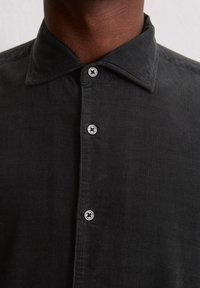 Gros plan sur le devant d'une chemise en velours côtelé noir avec un col et trois boutons en argent visibles sur le cou et le haut de la poitrine d'une personne.