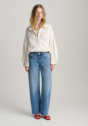 Nainen, jolla on päällään löysä valkoinen paita, korkeavyötäröiset leveälahkeiset siniset farkut, punaiset avoimet korkokengät ja hopeaketju kaulakoru, jossa on sydänriipus.