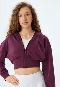 Sudadera corta morada con cierre de cremallera y cintura fruncida, que presenta mangas largas y una textura suave, usada sobre una camiseta de tirantes clara.