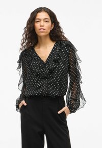 Blusa nera con pois, scollo a V con volant, maniche lunghe trasparenti e chiusura con bottoni. Abbinata a pantaloni neri. Tessuto leggero e testurizzato.