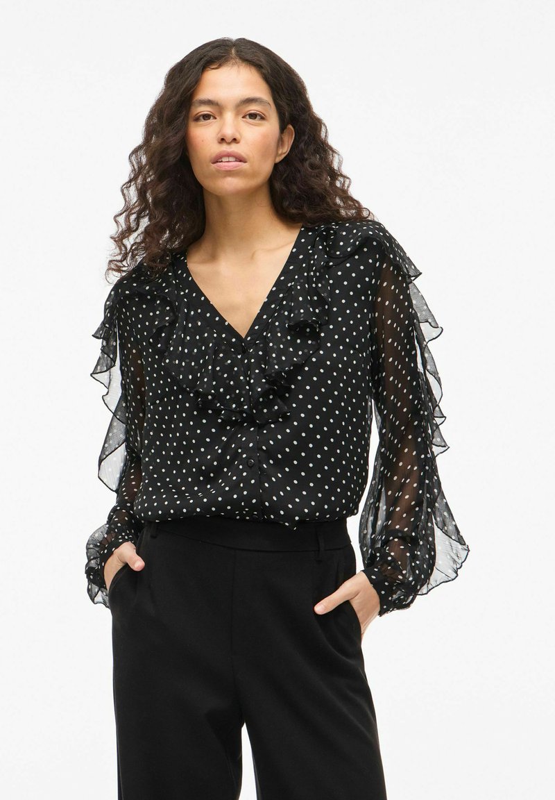 Blusa nera con pois, scollo a V con volant, maniche lunghe trasparenti e chiusura con bottoni. Abbinata a pantaloni neri. Tessuto leggero e testurizzato.