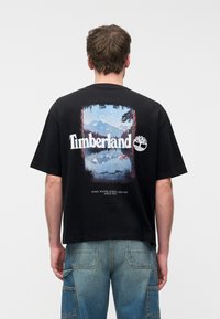 Άνδρας που φοράει μαύρο t-shirt Timberland με σχέδιο βουνού και λίμνης και μπλε cargo τζιν, όρθιος με την πλάτη προς τον θεατή σε λευκό φόντο.