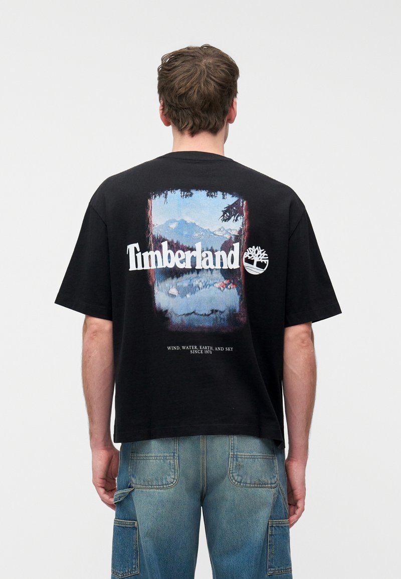 Άνδρας που φοράει μαύρο t-shirt Timberland με σχέδιο βουνού και λίμνης και μπλε cargo τζιν, όρθιος με την πλάτη προς τον θεατή σε λευκό φόντο.