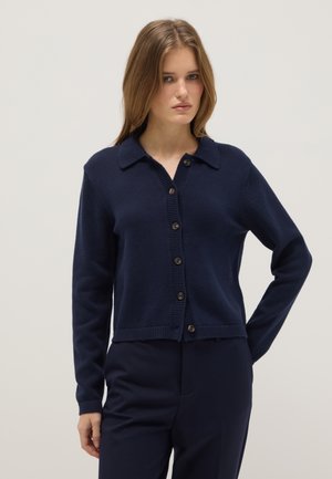 Vrouw met lichtbruin haar, gekleed in een marineblauwe cardigan met knopen en kraag, gecombineerd met een bijpassende marineblauwe broek, staand tegen een effen achtergrond.