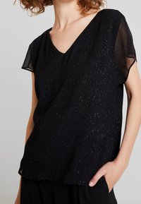 Black short-sleeve top with a V-neck, featuring a textured fabric with gold speckles and a relaxed fit. Accented with sheer sleeves.