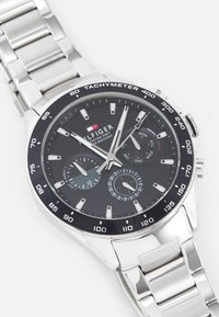 Montre en acier inoxydable argenté avec un cadran noir, aiguilles argentées, sous-cadrans pour la date et le jour, tachymètre sur la lunette et un bracelet à maillons.