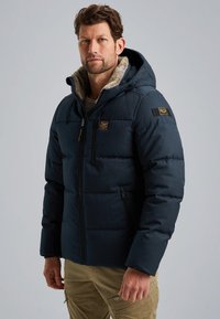 Muž v námořní modrej prešívanej bunde s kapucňou so syntetickým kožuchom a khaki cargo nohavicami, stojaci pred jednoduchým svetlosivým pozadím.