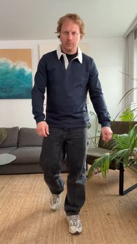 Nāvijas džemperis ar baltu apkakli, tumši džinsi un pelēki sporta apavi. Telpā ir augi un pelēks dīvāns, fonā redzama abstrakta sienas māksla.