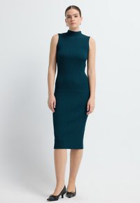 Ribbed, zielona sukienka do kolan z wysokim dekoltem i bez rękawów. Połączona z czarnymi, spiczastymi butami, które mają subtelny detal w postaci kokardy.