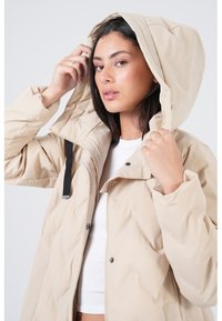 Beige gewatteerde jas met een capuchon, met een rits aan de voorkant en zwarte accenten. Gestructureerde stof met een gladde afwerking. Model houdt de capuchon vast.