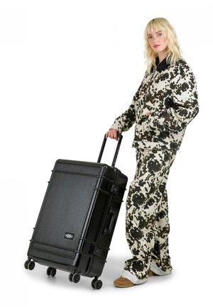 Valigia rigida nera con finitura strutturata, maniglie laterali, ruote rotolanti e serratura a combinazione; abbinata a un outfit beige e nero con stampa a motivo mucca.