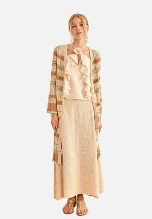 Mujer con blusa beige con volantes, falda larga texturizada, cárdigan de punto abierto a rayas y sandalias con cuentas, posando frente a un fondo blanco.