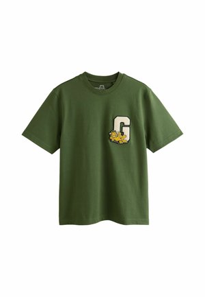 Mand iført mørke solbriller, grøn t-shirt med grafik, beige shorts og brune sko står på en grussti i en have.