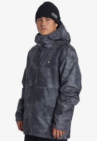 Šedá, strukturovaná bunda s kapucí a předním zipem. Má nášivku s diamantovým logem na hrudi a nastavitelné manžety. Nosí se s černou čepicí.