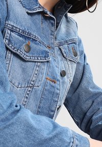 Ljusblå jeansjacka med krage, framfickor och svarta metallknappar. Tyget har en sliten yta och synliga sömmar.