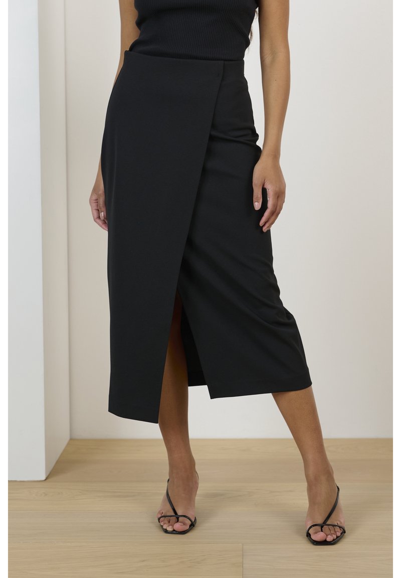 Svart omslagskjol i mjukt tyg, med en frontslits och en elegant design. Bärs med svarta sandaler med remmar på ett ljust trägolv.