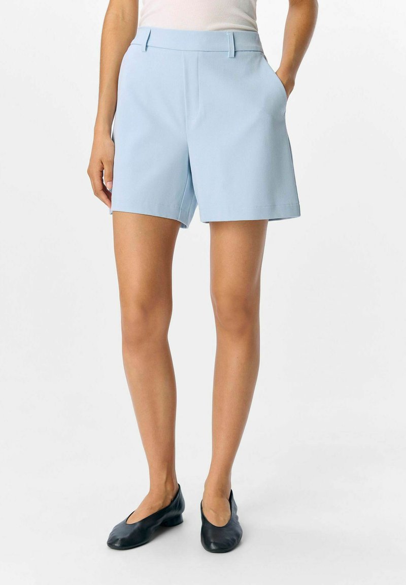 Pantaloni corti azzurri su misura con una trama liscia, dotati di tasche laterali e frontale piatto. Indossati con scarpe slip-on nere.