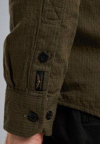 Camicia verde con bottoni, realizzata in tessuto testurizzato, caratterizzata da bottoni neri, un motivo cucito e un'etichetta con dettagli dorati.