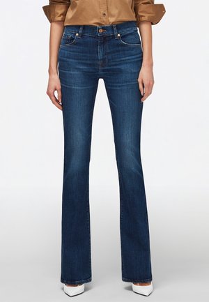 Frau trägt dunkelblaue Bootcut-Jeans, beige langärmliges Hemd mit hochgekrempelten Ärmeln und weiße spitze Schuhe, stehend vor weißem Hintergrund.