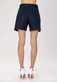 Shorts en coton bleu marine avec une coupe droite, une poche arrière avec détail de bouton et une taille plate, associés à des sandales à talons blancs.