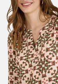 Blusa con estampado que presenta una base crema con diseños florales en verde y rosa, un escote en V y un delicado ribete de encaje a lo largo de la abertura.