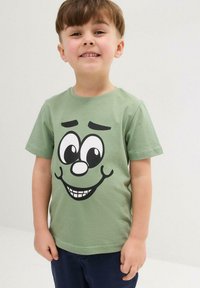 Maglietta in cotone verde con un grande volto fumettistico nero, caratterizzato da ampi occhi, un grande sorriso e sopracciglia, indossata con pantaloni scuri.