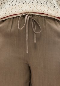 Μπεζ πλεκτό τοπ με μοτίβο διαμαντιού και κόκκινο περίγραμμα πάνω από χαλαρά, καφέ παντελόνια με κορδόνι στη μέση.